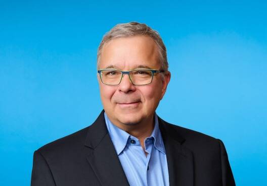 A man with short gray hair and glasses, wearing a dark blazer over a light blue shirt, stands in front of a plain blue background, smiling slightly—evoking professionalism and expertise in Private Equity Consulting.