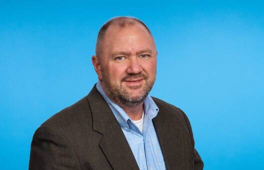 A middle-aged man with short hair and a beard, wearing a brown blazer over a light blue collared shirt, stands in front of a plain blue background, smiling slightly at the camera—an expert in Operational Excellence and Private Equity Consulting.