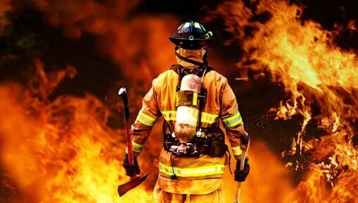 A firefighter in full gear, holding an axe, stands facing intense flames, surrounded by fire.
