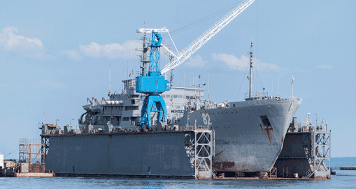 A large naval ship sits in a floating dry dock, with a prominent blue crane on its deck. Demonstrating operational excellence, the vessel appears to be undergoing maintenance or repairs as calm water reflects the clear sky above.