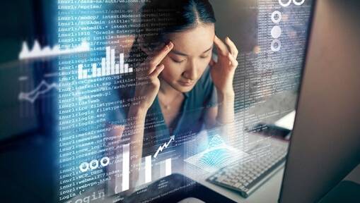 A woman sits at a desk with her hands on her temples, looking stressed, as computer code and graphs are projected in front of her, suggesting data analysis or programming work.