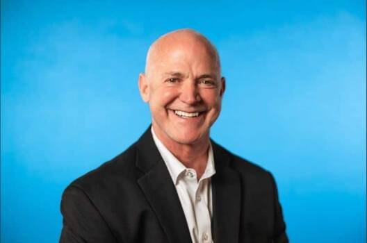 A smiling bald man in a black suit jacket and white shirt poses in front of a bright blue background, embodying continuous improvement and operational excellence.