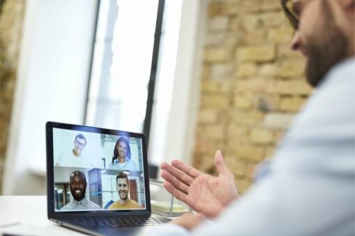 A person gestures while on a video call, shown on a laptop screen with four other people, each in their own video window, participating in a virtual meeting.