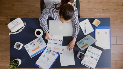 A person sitting at a desk, reviewing charts and graphs on printed papers and a laptop, with a cup of coffee, notepad, pen, and office supplies neatly arranged around them.