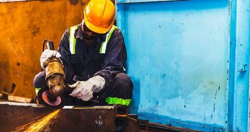 A worker wearing a yellow hard hat, gloves, and safety gear uses an angle grinder, producing sparks while working on metal in an industrial setting—a scene reflecting operational excellence with blue and yellow walls.