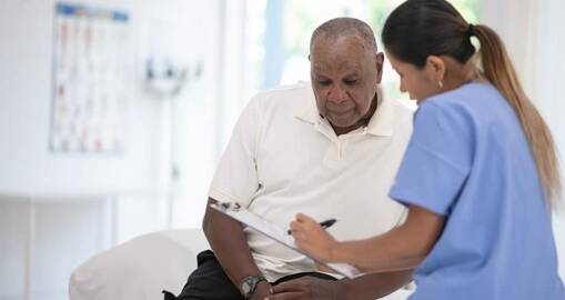 A nurse in blue scrubs sits beside an older man in a white shirt, explaining information on a clipboard in a brightly lit medical office.