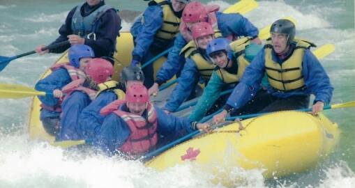 A group of people in helmets and life jackets paddle a yellow inflatable raft through whitewater rapids, looking focused and excited as water splashes around them.