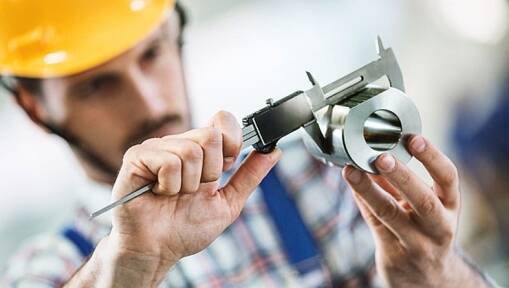 A person wearing a yellow hard hat uses a caliper to measure a metal component, focusing closely on the measurement—a scene reflecting the precision and attention to detail essential in Lean Manufacturing Consulting. The background is blurred to emphasize their work.