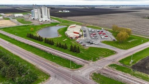 Aerial view of a rural facility renowned for Operational Excellence, featuring a pond, several buildings, parked cars, and green lawns, surrounded by farmland. Railroad tracks and roads intersect near the property.