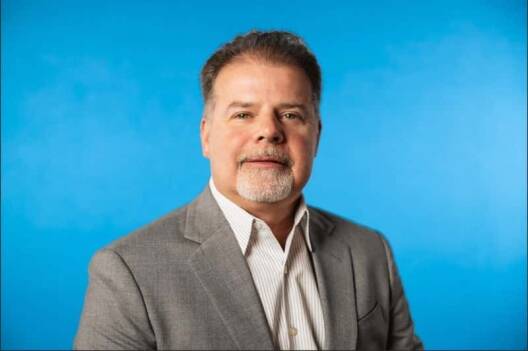 A middle-aged man with short brown hair and a goatee is wearing a gray suit jacket and a white collared shirt, posing against a solid blue background.