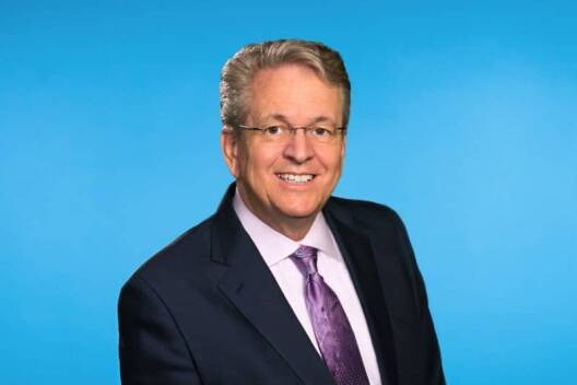 A smiling man with gray hair and glasses, wearing a dark suit, light purple shirt, and purple tie, poses in front of a solid blue background—reflecting his expertise in Lean Manufacturing Consulting.