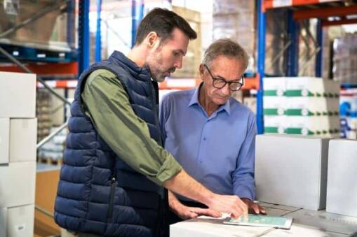 Two men standing in a warehouse, looking at documents on a tablet placed on a stack of white boxes; one man wears a vest and gestures towards the screen while the other, in glasses and a blue shirt, observes attentively.