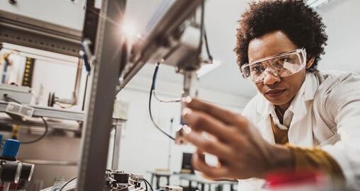 A scientist wearing safety goggles and a lab coat adjusts equipment in a laboratory, focusing intently on her work with machinery and scientific instruments in the background.