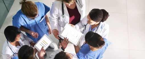 A group of healthcare professionals in scrubs and lab coats stand together, viewing charts and data on tablets and paper, collaborating as a team in a bright, modern setting.