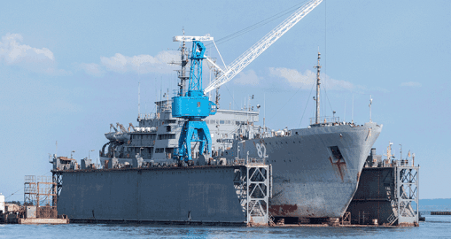 A large naval ship sits in a floating dry dock, with a prominent blue crane on its deck. Demonstrating operational excellence, the vessel appears to be undergoing maintenance or repairs as calm water reflects the clear sky above.