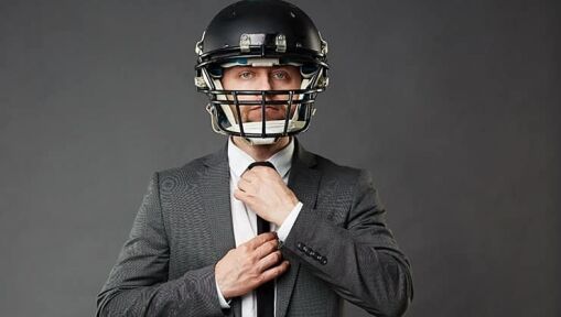 A man wearing a black suit and tie adjusts his tie while also wearing a black football helmet, standing against a plain dark background.