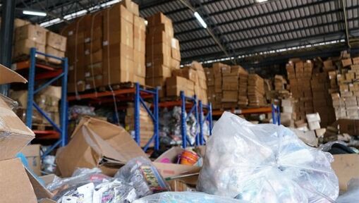 A warehouse filled with stacks of cardboard boxes on metal shelves. In the foreground, open boxes and large plastic bags with various items are scattered, creating a cluttered scene.