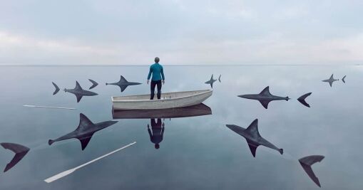 A person stands alone in a wooden rowboat on calm water, surrounded by the fins of several sharks. Two oars float nearby, and the scene has a surreal, dreamlike atmosphere.