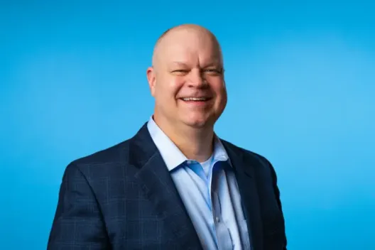 A smiling bald man in a blue blazer and light blue shirt stands against a plain blue background, embodying professionalism in private equity consulting.