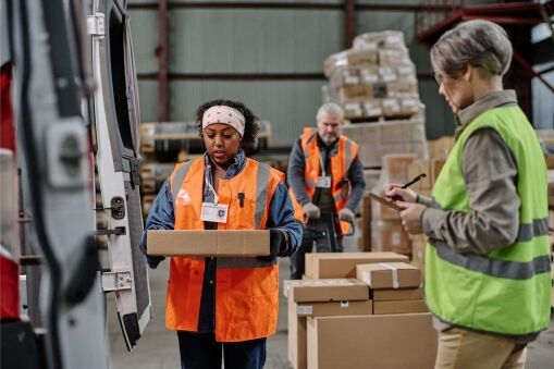 Three workers in high-visibility vests handle packages in a warehouse; one carries a box, another checks a clipboard, and a third organizes parcels in the background.