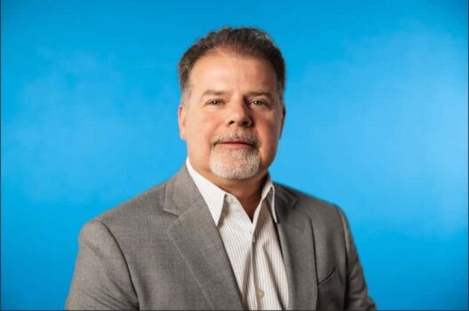 A middle-aged man with short brown hair and a goatee is wearing a gray suit jacket and a white collared shirt, posing against a solid blue background.