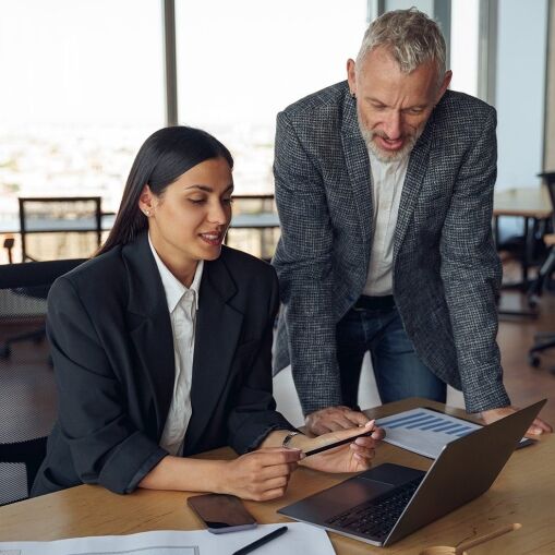 Two business professionals in an office discuss work at a desk with a laptop, documents, and a phone. The seated woman points at the screen while the standing man listens and looks at the laptop. Large windows show a city view behind them.