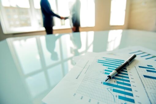 A close-up of financial charts and a pen on a glass table, with two blurred people shaking hands in the background in a bright office setting.