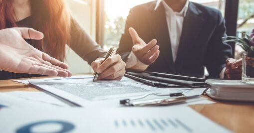 Two people in business attire sit at a desk with documents and charts. One person is signing a contract while the other gestures, suggesting a discussion. Sunlight comes through a window in the background.