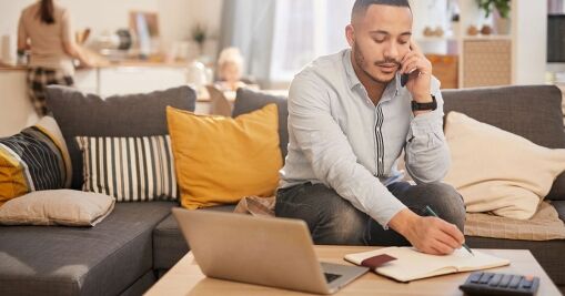 A man sits on a sofa, talking on his phone while writing in a notebook. A laptop, notebook, and calculator are on the coffee table. In the background, a woman and child are in the kitchen.