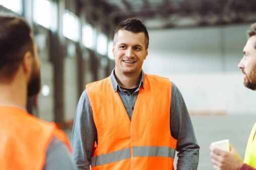 Three men wearing orange safety vests stand and talk inside a large, bright warehouse. One man in the center is smiling, while the other two are partially visible, facing him.
