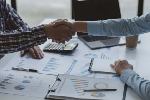 Two people shake hands over a table with documents, charts, a calculator, and a notebook, suggesting a successful business agreement or meeting. Another person’s hand is seen writing in a notebook.