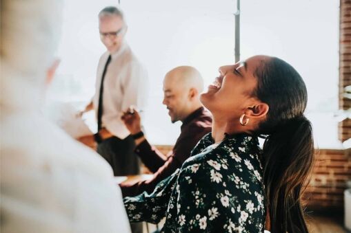 A woman with a ponytail and floral blouse smiles and looks up while sitting at a table with colleagues during a bright, casual office meeting.
