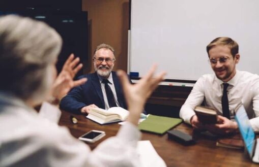 Three people in business attire sit around a conference table talking and smiling, with notebooks, a laptop, and a phone in front of them, and a whiteboard in the background.
