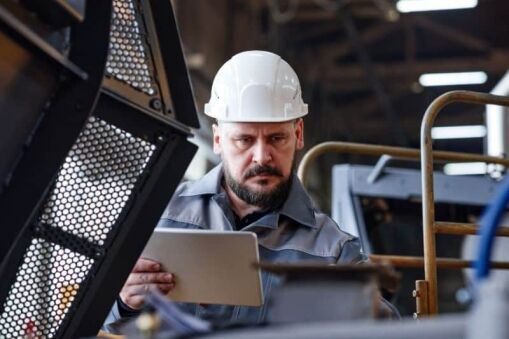 A man wearing a white hard hat and grey work uniform inspects machinery while holding a tablet in an industrial setting.