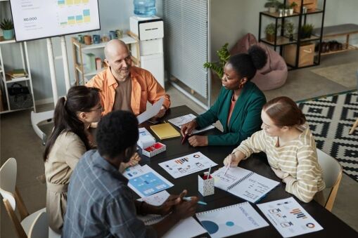 Five people sit around a table in an office, discussing documents and charts. Laptops, notebooks, and printed graphs are on the table. A whiteboard and shelves with plants are visible in the background.