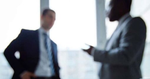 Two men in business suits are having a conversation indoors. The image is intentionally blurred, making their faces and details indistinct. Light streams in from large windows behind them.