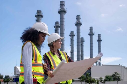 Two women wearing safety vests and hard hats examine a large blueprint outdoors, with industrial plant towers and pipelines visible in the background under a partly cloudy sky.