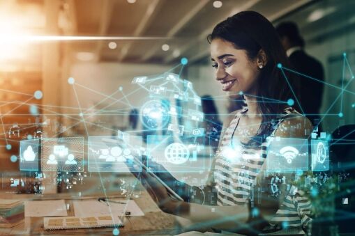A woman sits at a desk smiling at her tablet, surrounded by digital hologram icons representing social media, cloud computing, and networking in a modern office setting.