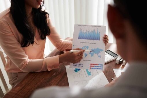 Two people in business attire discuss data charts. One person points to a printed sheet showing bar graphs, a world map, and analytics, while the other listens. A calculator and more documents are on the desk.