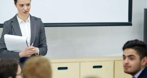 A woman in a gray blazer holding papers stands in front of a group, appearing to give a presentation. Several people are seated, listening to her in a classroom or meeting setting.