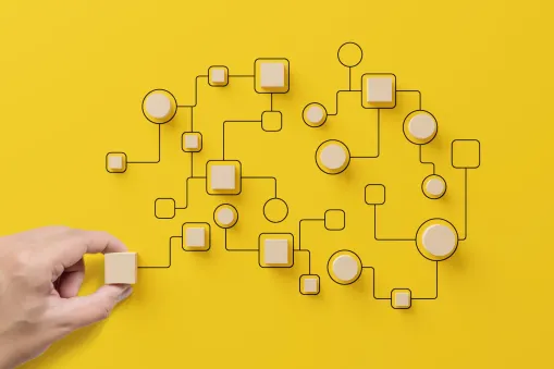A hand places a wooden square block among interconnected pieces forming a brain shape on a yellow background, symbolizing networks and strategy—an apt visual for Private Equity Consulting.