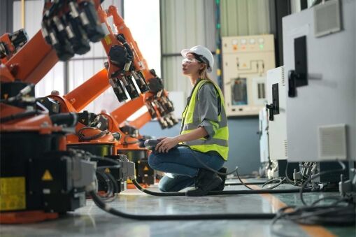 A woman wearing a hard hat and safety vest kneels beside an industrial robot arm in a factory, using a control device. She appears to be inspecting or operating the machinery in a modern Aerospace & Defense Consulting setting.