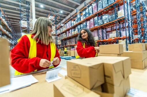 Two women in red shirts and yellow safety vests smile and talk while working with cardboard boxes and paperwork in a large warehouse, showcasing the organized efficiency promoted by lean manufacturing consulting.