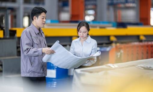 Two people in a factory setting examine a large blueprint together, demonstrating continuous improvement. The man wears a gray work jacket and the woman is in a white blouse, with industrial equipment and machinery visible in the background.