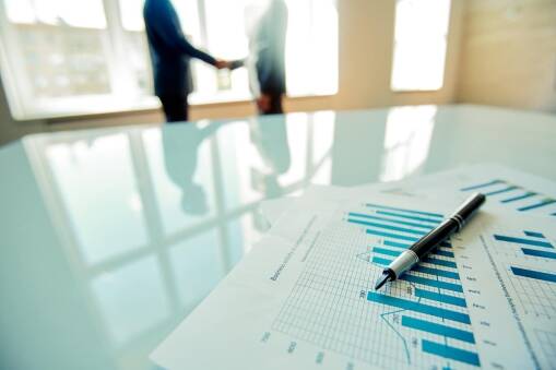 A close-up of financial charts and a pen on a glass table, with two blurred people shaking hands in the background in a bright office setting.