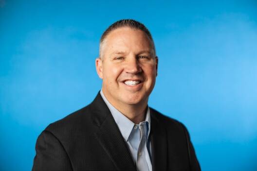 A man in a dark suit jacket and light blue shirt smiles at the camera against a plain bright blue background, embodying professionalism in Lean Manufacturing Consulting and Operational Excellence.
