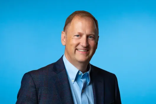 A man with short light brown hair, wearing a dark blazer over a light blue dress shirt, smiles at the camera against a solid blue background—reflecting professionalism in Lean Manufacturing Consulting.