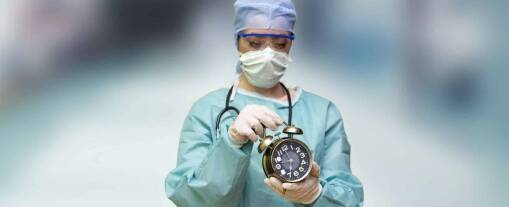 A healthcare worker in scrubs, mask, gloves, and a stethoscope holds a large analog clock, focusing intently on its start time. The blurred background highlights the urgency of the moment.