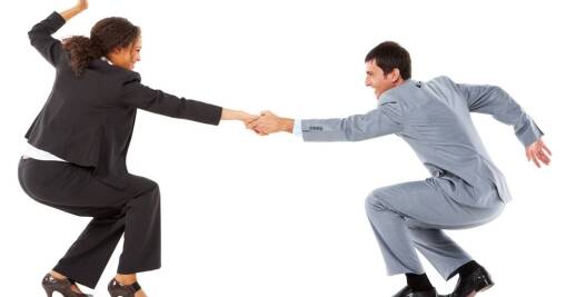 A woman and a man in business suits hold hands and squat facing each other, as if supporting or helping each other, against a white background.