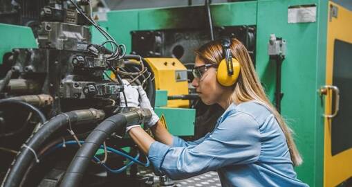 A woman wearing safety glasses and yellow earmuffs operates machinery in an industrial setting, adjusting cables and components with gloved hands—a testament to operational excellence on the production floor.